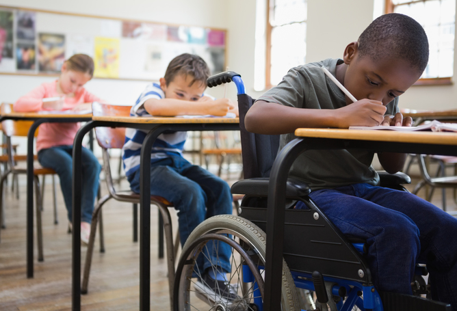 A picture of three young students in a classroom with one of them on a wheelchair