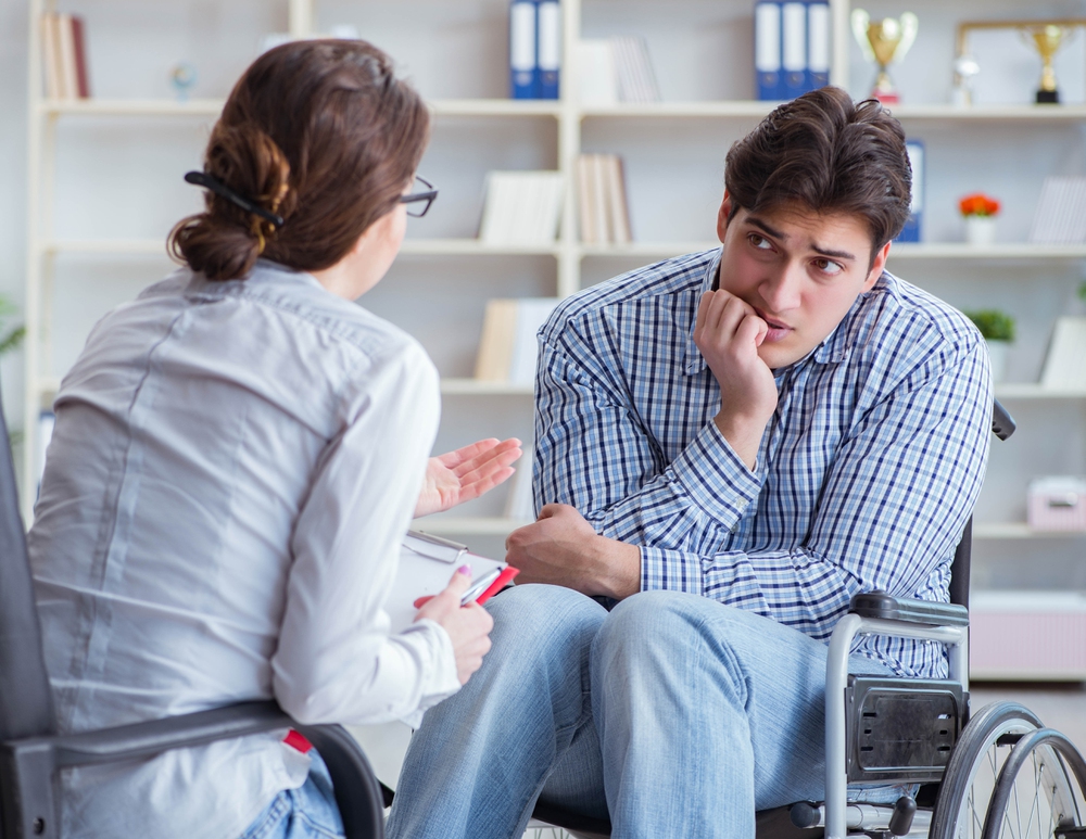 A woman speaks to a man with disability in a wheelchair during a therapy session in a bright, well-organized office. Books and trophies are in the background.