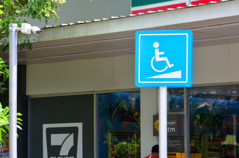 Blue accessibility sign with a wheelchair symbol indicates a ramp, positioned outside a store beneath a security camera. Signage promotes inclusivity.