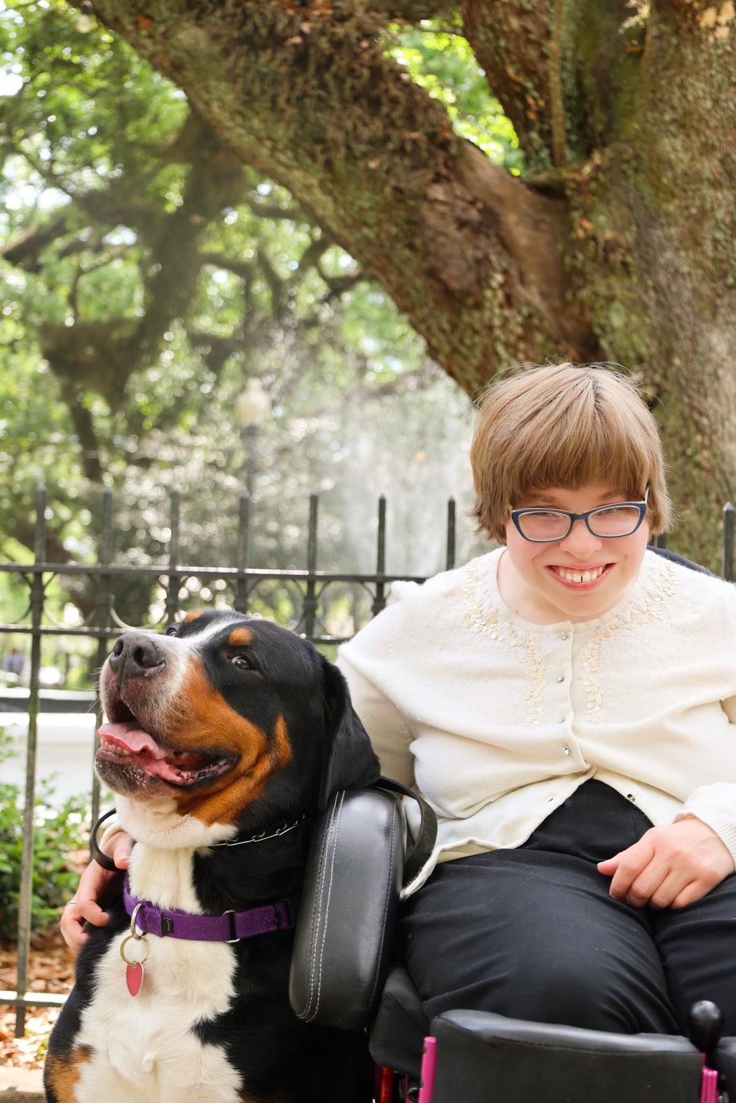A person in a white sweater smiles joyfully, sitting in a wheelchair beside a large, friendly dog with a purple collar. They are under a tree in a park setting.
