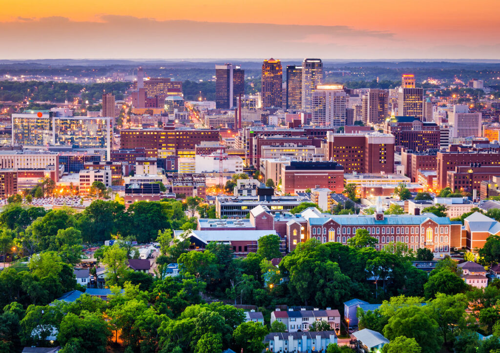 Aerial view of Alabama at sunset. The skyline features illuminated skyscrapers with a foreground of lush greenery and residential buildings.