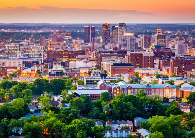 Aerial view of Alabama at sunset. The skyline features illuminated skyscrapers with a foreground of lush greenery and residential buildings.