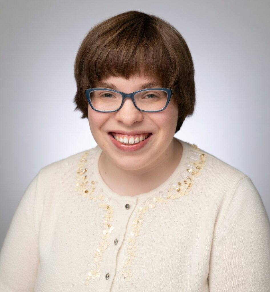 This is Addie, smiling with short brown hair and glasses, smiling warmly, wearing a cream top with decorative sequins. The background is a soft gray color.
