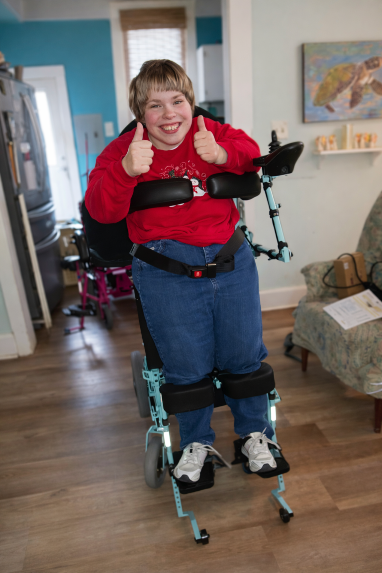 This is Addie in a red sweater gives a thumbs up while standing supported by an Automatic Wheel chair in a cozy, welcoming room with cheerful decor.